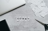 Black and white photo of a desk. The letters “SORRY” have been individually cut out and placed in the middle. There is a pair of scissors, paper scraps, a laptop, and a book around the letters.