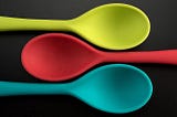 Three multi-colored plastic spoons posing on top of  a dark surface.