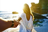 back view of woman with long brown hair wearing white dress walking on rocky beach holding man’s hand with arm outstretched behind her