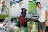 Two young adults pose in front of recycling waste bins.