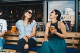 Two Smiling Women Sitting on Wooden Bench Drinking a Beer