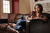 Black woman with locs wearing chocolate brown shirt and jeans curled up writing in a journal on a brown leather couch.