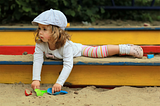 A young girl playing in a colorful wooden sandbox with colorful shovels.