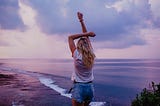 Woman stretching with her arm up in front of a purple hued ocean and sky