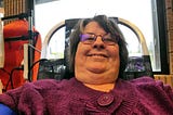 A smiling person with purple glasses is taking a selfie while reclining in a chair, possibly during a blood donation procedure. Behind them, medical equipment can be seen, which suggests a healthcare setting.