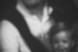 A blurred, black and white photo of a white man with dark hair, white collard shirt and a dark sweater wrapped around his neck. He holds a small white child in a white sleeveless dress with dark straps. Through the blur, they both seem to be smiling.