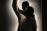 A photograph of a Black teen standing in front of a silhouette of a slightly taller Black man. Both the teen and the silhouette hold one raised fist in the air. The silhouette is outlined by white light. It is too dark to see what the teen is wearing. The teen’s body faces the viewer and he looks to the right, towards his raised fist.