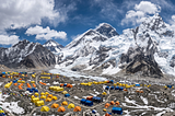 A mountaineering base camp on rocky glacial ground, with small tents dwarfed by a vast snow-covered peak rising into the sky above them.