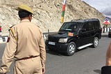 The Dalai Lama is waving his right hand in black SUV. The SUV has a small prayer flag with multi color stripes in front. There is a policeman standing in front to manage the crowds.
