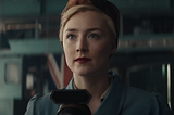 Close-up of a woman in attire, holding a microphone, in a factory-like setting with a British flag in the background.