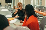 A Black Woman with strawberry blonde hair in sitting at a board room table signs to a South Asian woman with black hair wearing orange