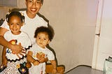 A young Alexsis, one of her brothers, and their mother standing in a kitchen. Their mother holds the children from behind as they all smile for the camera. There’s a birthday cake on the table in front of them.