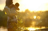 A brown-haired woman cuddles an infant, and they look out together at body of water at dusk.