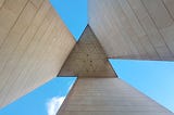 Looking up from beneath the National Carillon Monument against a blue sky in Canberra, Australia. The geometry of the monument makes a equilateral triangle in the middle, with cement slabs and the bright blue sky creating paired beams come from all three sides of the triangle.