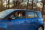 An obese white woman with short brown hair and a teal shirt sits behind the wheel of a blue and black hatchback vehicle.