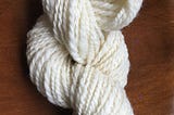 a twisted skein of undyed, overspun 2-ply yarn on a wooden surface.