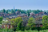 Terraces of houses near the city centre of Stoke on Trent, between lines of green-leaved trees. In the background, a church steeple rises above the treeline into a cloudless sky.