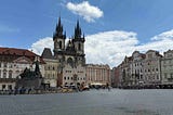 A large historic square in Prague with cobblestone paving, people walking, and outdoor seating. The Gothic Church of Our Lady before Týn rises in the background with its two tall spires, and the Jan Hus Memorial stands prominently in the center, surrounded by ornate buildings with red‑tiled roofs under a partly cloudy sky.