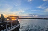 A pontoon is leaving the frame of the photo in the bottom left corner of the photo. You can see two people on the boat. A forested shoreline is in the distance and the sun is descending into the horizon behind the boat.