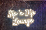 Photo of a neon sign, “Sip ‘n Dip Lounge” hanging on a chain on a rattan wall