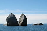 Photo of the split apple rock off the coast of ateroa/New Zealand https://en.wikipedia.org/wiki/Split_Apple_Rock The rock is large, round and grey and split in two. It is surrounded by blue water. The background has white clouds low but clear sky up above. There is a bird sittingon the right hand half of the rock.