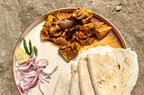 In a plate, a flatbread along with meat curry is served with sliced onions, lemon, and green chillies on the side.