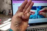 A close-up photo shows a person’s hand making a sign with their thumb and little finger touching. The other three fingers are extended straight up. In the background, there’s a blurred view of a laptop screen displaying what appears to be a webpage.