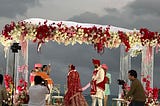 An Indian Bride and Groom are shown going in circle around a fire under a canopy decorated with flowers.