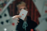A blurred figure holding the ace of diamonds between their fingers at the viewer, surrounded by a whirling ring of playing cards