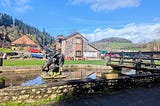 A winged metal dragon sculpture stands in a small pond beside a stone cottage and wooden footbridge, with green hills and buildings near Tintern Abbey under a bright blue sky.