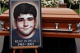 A picture of a chubby, dark haired 20-year-old male sits in front of a closed casket. The picture says “Rest in Peace 1983–2003.”