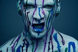 A growling man painted gray with drips of blue and red paint running down his face and out of his mouth
