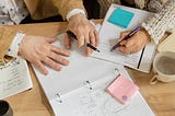 Two people’s hands holding pens on a desk over a binder, several post it notes and a notebook
