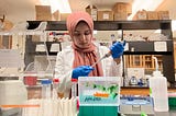 Khloud wearing a white lab coat and a pink scarf and working with scientific tools in a lab.