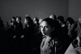 Black and white photo with a young girl in focus glancing toward the camera. Her face is absent of emotion. No smile, no frown either. The room is crowded with people. She looks alone and maybe afraid.