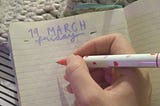 My hand holds a pen on top of my journal page. My journal page has the heading 19th March, Friday, on it.