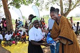 A young black girl wearing a white t-shirt with the UN Trust Fund logo, a green headband and black skirt, is seen smiling receiving an award from a young black women wearing a yellow and black dress. In the background we can see people sitting and standing outside.
