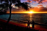 Beautiful brightly colored orange, yellow, purple, blue ocean beach sunset with black silhouettes of a palm tree and a couple (man & woman) holding hands in the suns reflection on the wet beach sand.