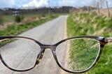 A view of the blue sky and road ahead, through a pair of rain-speckled eyeglasses.