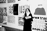 Photography of Sister Corita Kent stands in front of her work, including for Eleanor, at Immaculate Heart College in 1964.