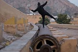 The back of a person in black clothing, and black hat, balancing on one leg on a large pipe while facing a mountain in the distance.