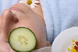 photo of woman with cucumber on eyes and flower on chest wearing blue robe; photo on Dr. James Goydos 2021 article on skin care self care and skin cancer prevention