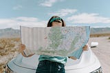 Woman sitting on car bonnet looking at map held in both hands hiding her face