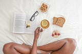 Woman sitting on a bed holding a cup of tea, with a book, glasses and a healthy breakfast in front of her.