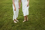 From the chest down we see two women in flowing white dresses standing on a field of green grass, holding hands.