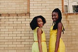 Two young black girls in yellow dresses, smiling, looking toward the photographer, standing in front of a brick wall.