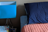 A side table stacked with books and a turquoise lamp, and a glimpse of my bed with a navy pillow case and salmon comforter.