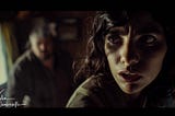 A tense moment in a dimly lit trailer where a young woman, Maya, stands defiantly, her expression a complex mix of fear and resolve. In the blurred background, a man stumbles, his face etched with shock. The charged atmosphere is palpable, reflecting a pivotal confrontation amidst a brewing storm.