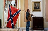 An often-republished photo of a January 6 rioter standing in the Capitol Building, holding a large confederate flag.