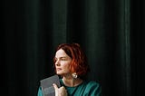 Woman with red hair holds a closed book and looks out of a window